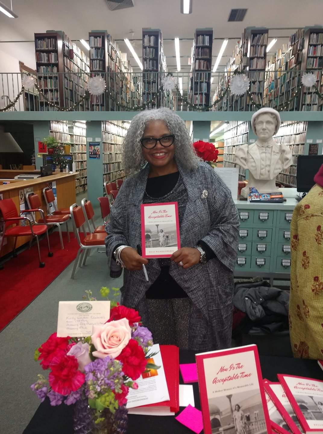 Kelly Walker Edwards holding up a copy of Now Is the Acceptable Time in a library reading room, flowers and more copies on the table