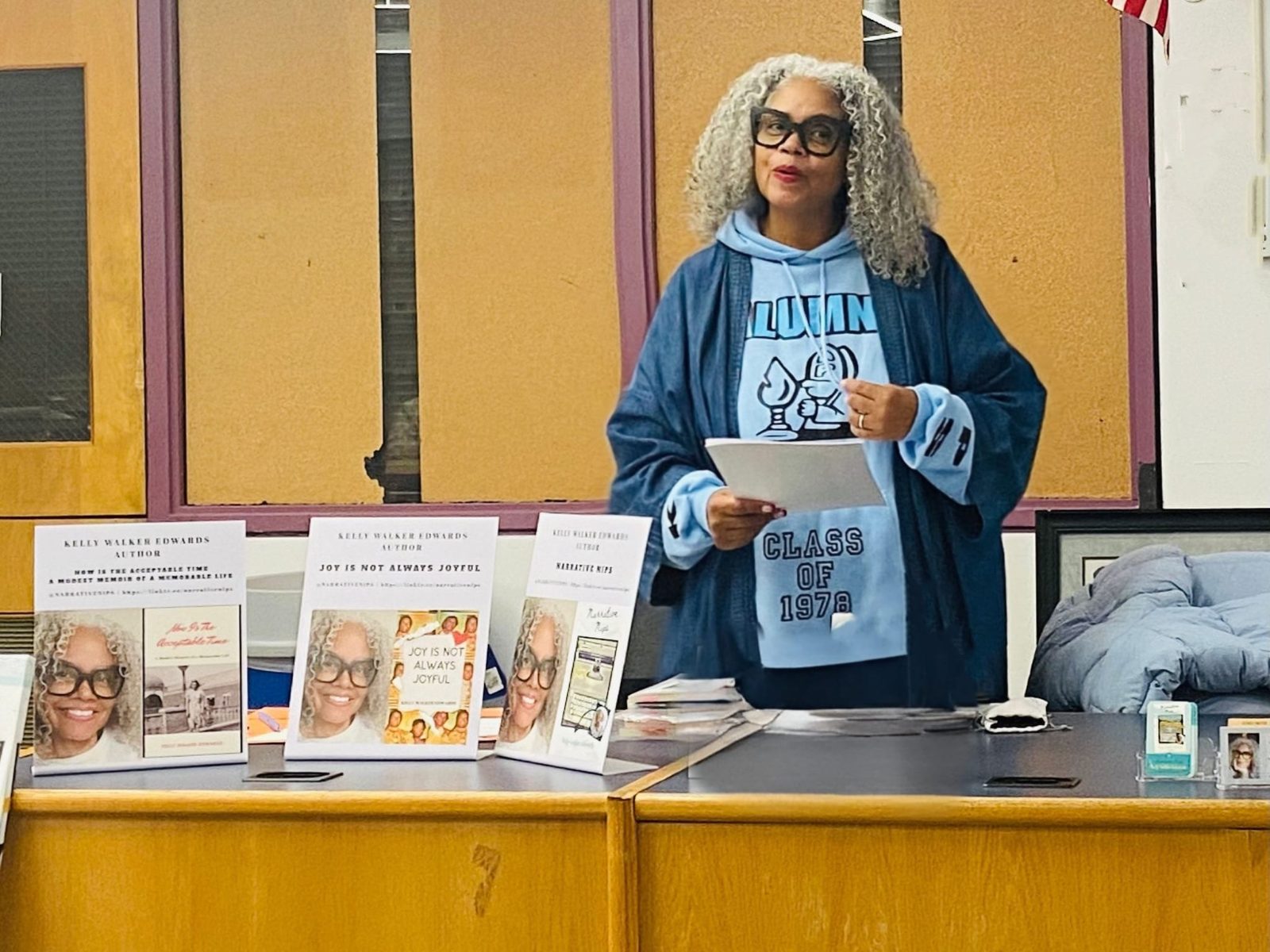 Kelly Walker Edwards reading from a lectern in an Asbury Park Class of 1978 alumni hoodie, her books displayed in front of her