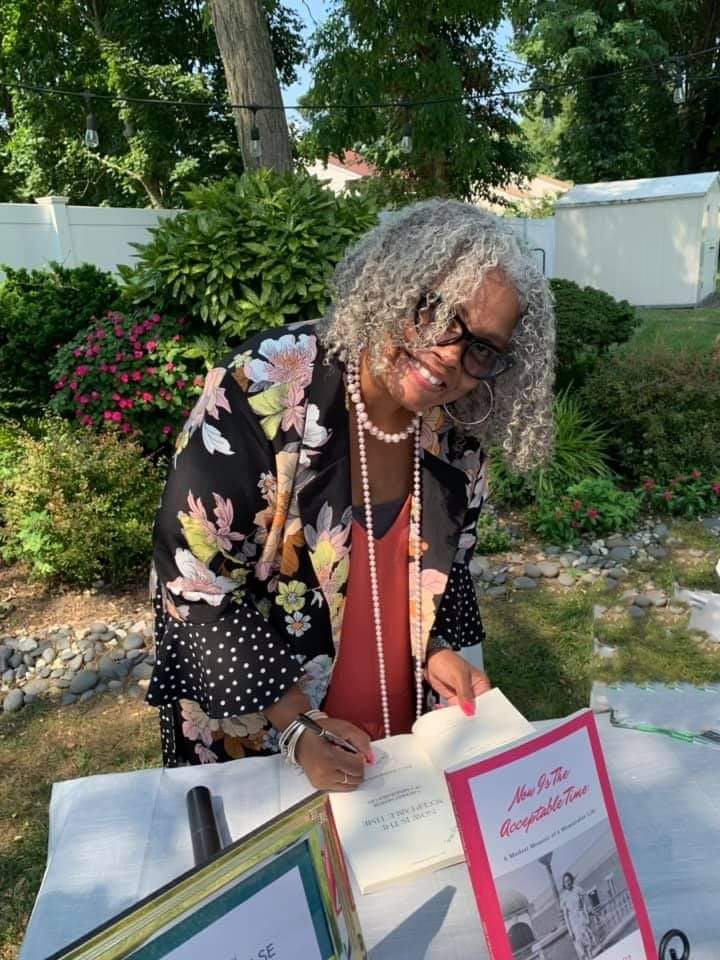 Kelly Walker Edwards signing a copy at a table outdoors, a garden and flowering shrubs behind her